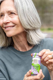 An older woman with gray hair smiles outdoors while holding a purple and green tube of Good Flower Farm Lavender Tea Tree Deodorant in a plastic-free tube.