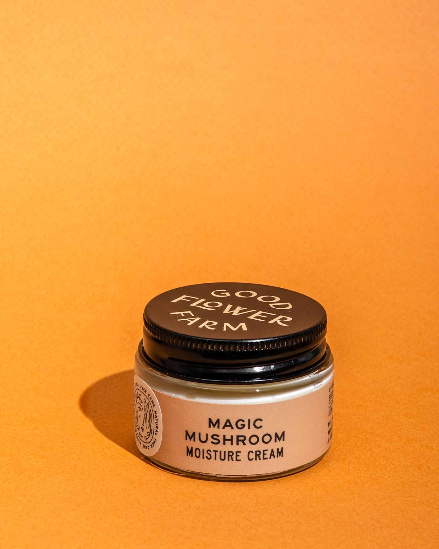 A small Good Flower Farm Magic Mushroom Moisture Cream jar with a black lid and beige label sits on an orange background. The cream is infused with adaptogenic maitake.