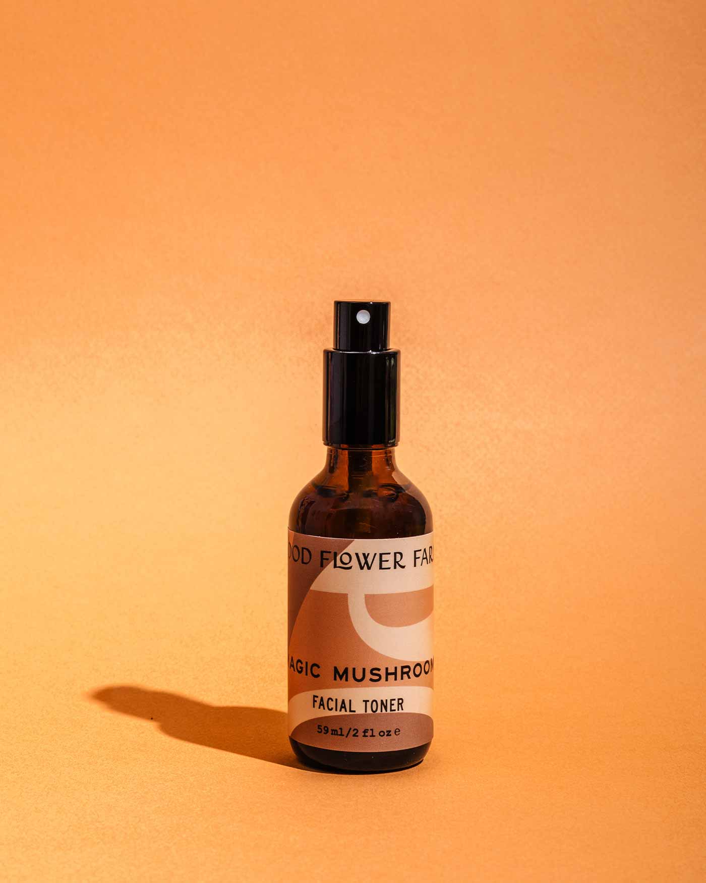 A brown glass spray bottle of Good Flower Farms Magic Mushroom Facial Toner stands upright on an orange surface, providing elemental protection and skin hydration, with a soft shadow beside it.