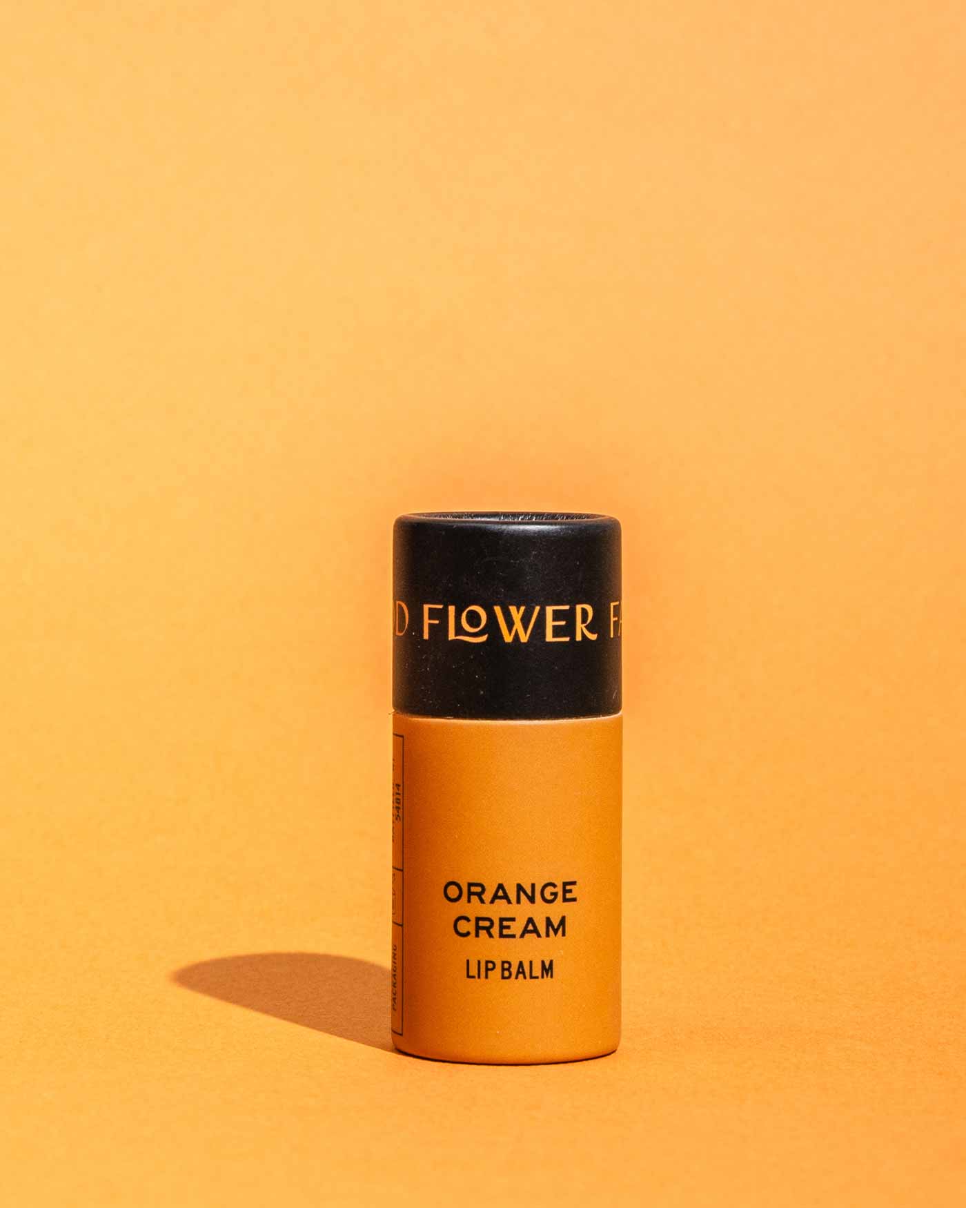 A cylindrical orange container of Good Flower Farms TESTER — Orange Cream Organic Lip Balm stands upright against an orange background, casting a small shadow to the left. Its top is black with gold text.