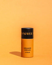 A cylindrical orange container of Good Flower Farms TESTER — Orange Cream Organic Lip Balm stands upright against an orange background, casting a small shadow to the left. Its top is black with gold text.