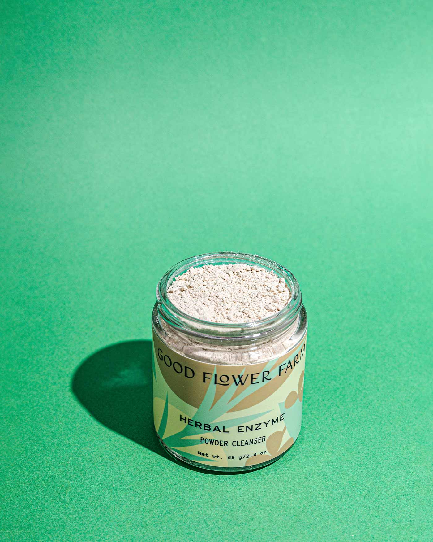 An open glass jar of Good Flower Farm Herbal Enzyme Powder Facial Cleanser sits on a bright green background, revealing its light powder packed with pineapple and papaya enzymes.