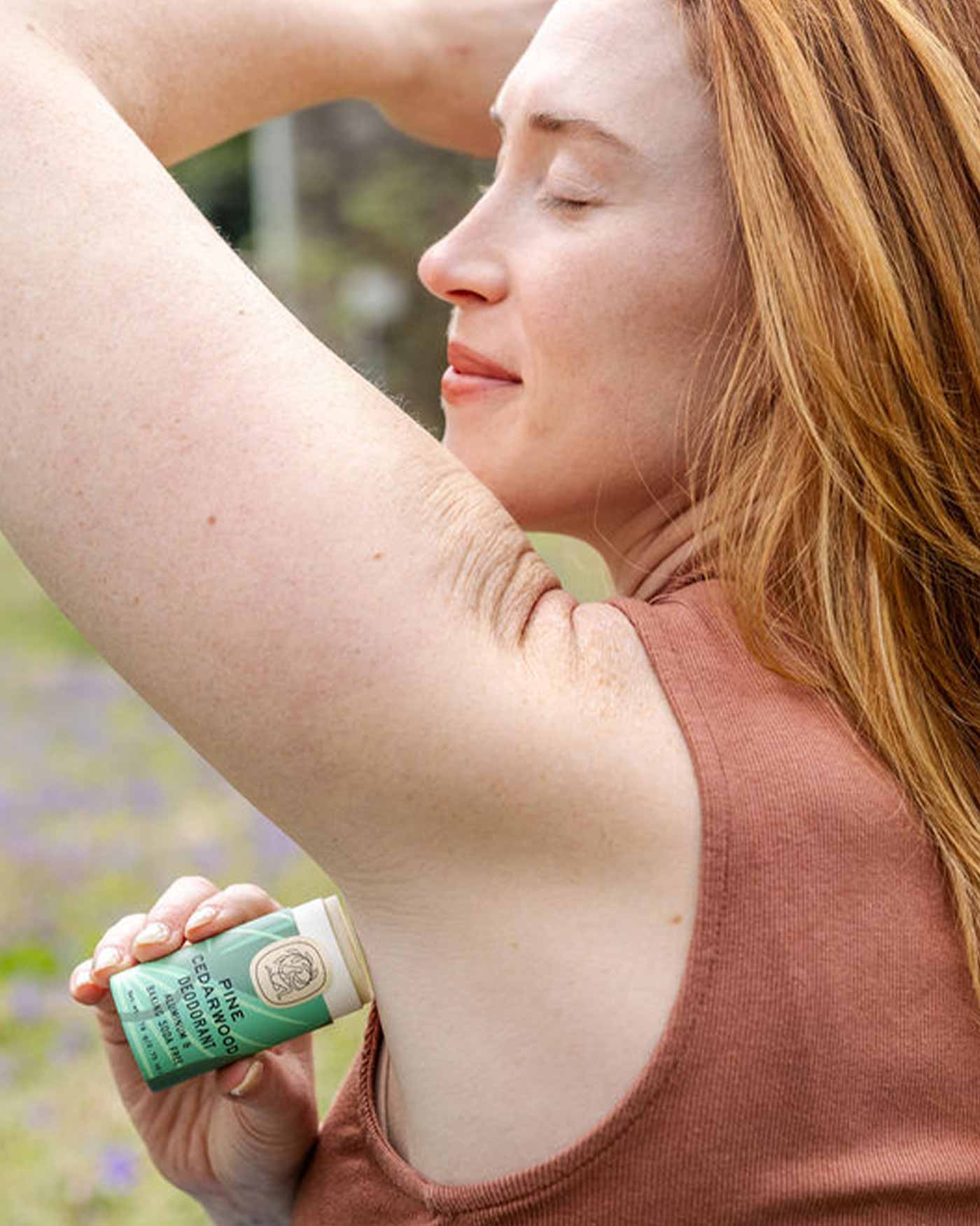 A red-haired woman in a sleeveless brown top relaxes outdoors with her eyes closed as she applies Good Flower Farms Pine Cedarwood Deodorant Plastic-Free Tube to her underarm.