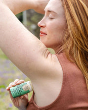 A red-haired woman in a sleeveless brown top relaxes outdoors with her eyes closed as she applies Good Flower Farms Pine Cedarwood Deodorant Plastic-Free Tube to her underarm.