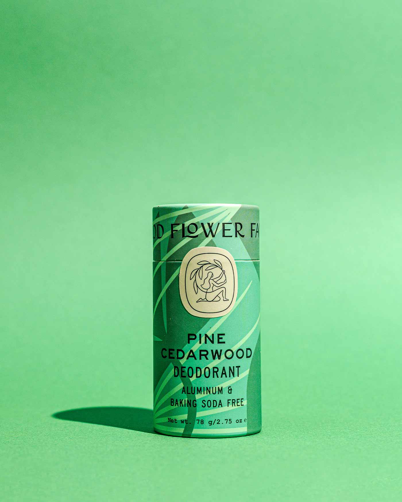 A green cylindrical tube of Good Flower Farm Pine Cedarwood Deodorant Plastic-Free stands upright on a matching background. The label notes its aluminum- and baking soda-free, in biodegradable packaging.