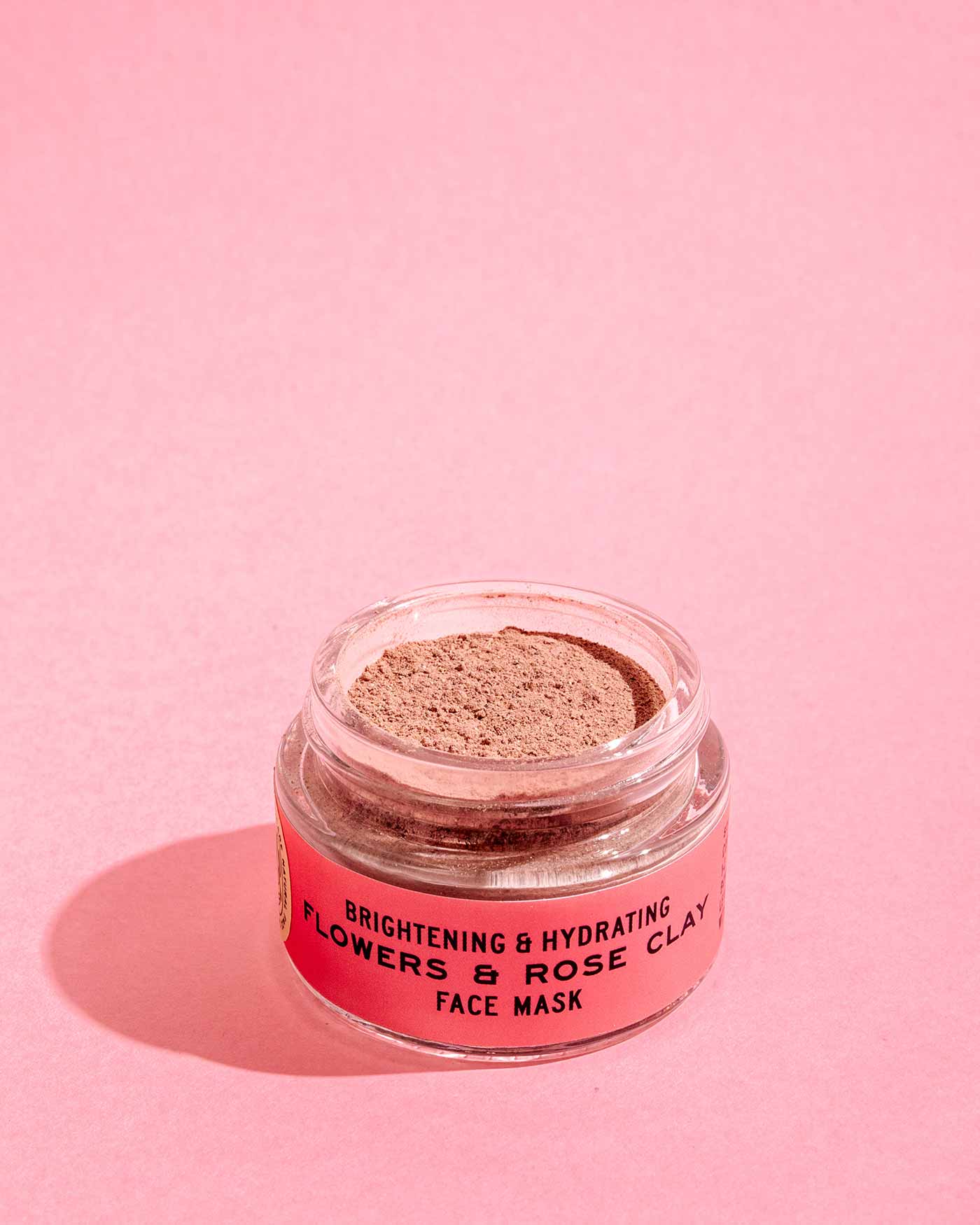 A clear jar of Good Flower Farm’s Flowers & Rose Clay Botanical Face Mask, with light brown powder and a pink label, rests on a pink surface, showcasing its brightening and hydrating floral blend against a matching background.