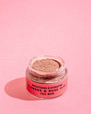 A clear jar of Good Flower Farm’s Flowers & Rose Clay Botanical Face Mask, with light brown powder and a pink label, rests on a pink surface, showcasing its brightening and hydrating floral blend against a matching background.