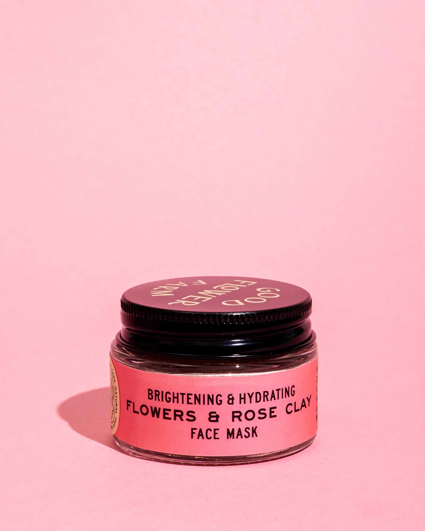 A small jar of Good Flower Farm Flowers & Rose Clay Botanical Face Mask with a black lid is set against a solid pink background.