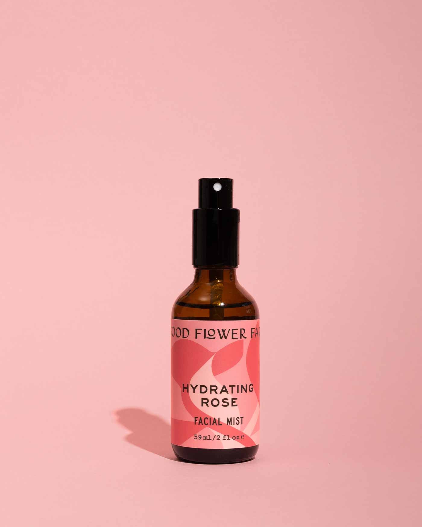 A brown glass bottle of Good Flower Farms Hydrating Rose Facial Mist stands on a pink surface, featuring rose hydrosol and aloe vera for deep hydration. The solid pink background casts a gentle shadow beside the bottle.