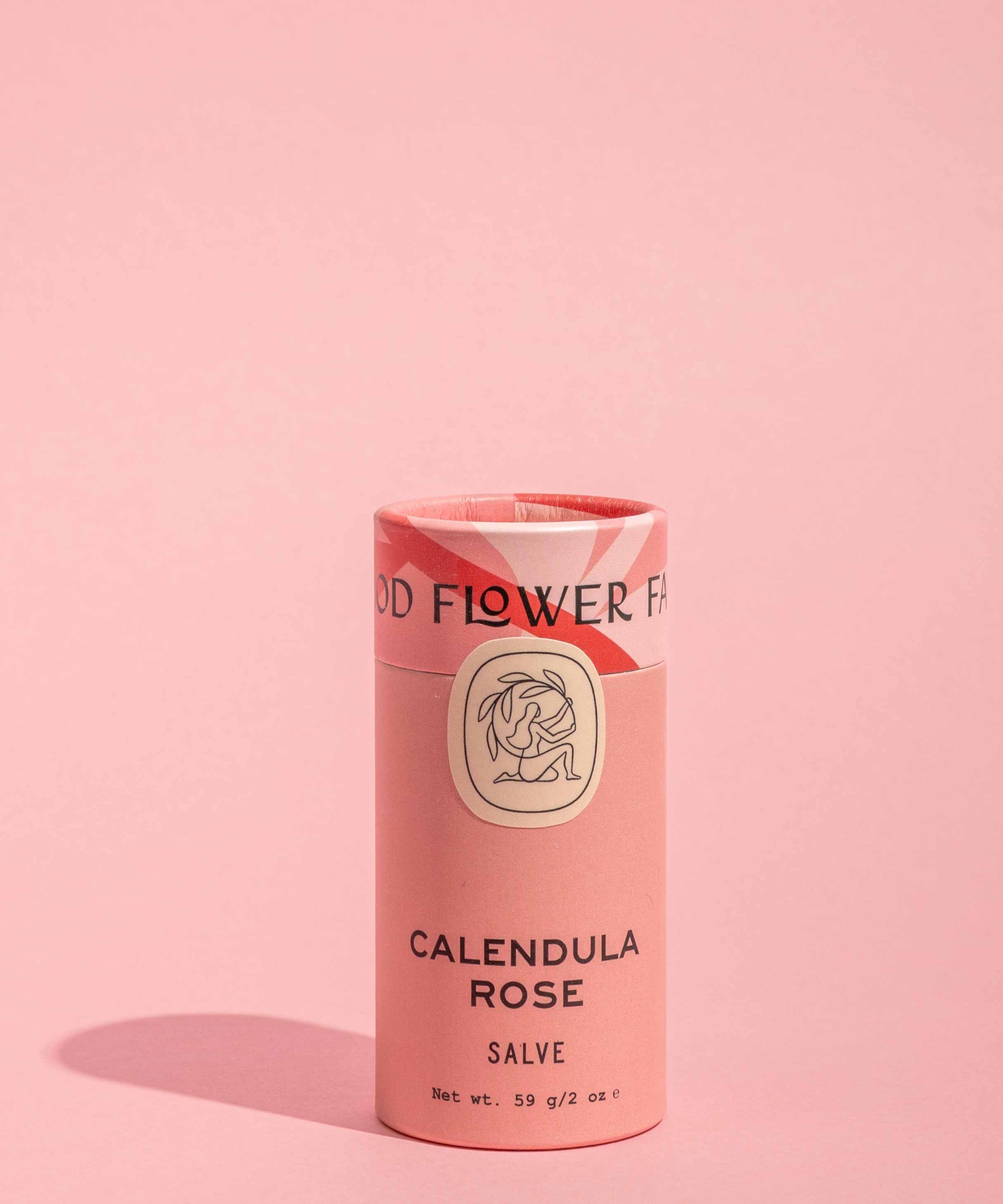 A pink cylindrical container of Good Flower Farm Calendula Rose Salve stands upright on a matching background, featuring simple black text, a floral illustration, and eco-friendly packaging.
