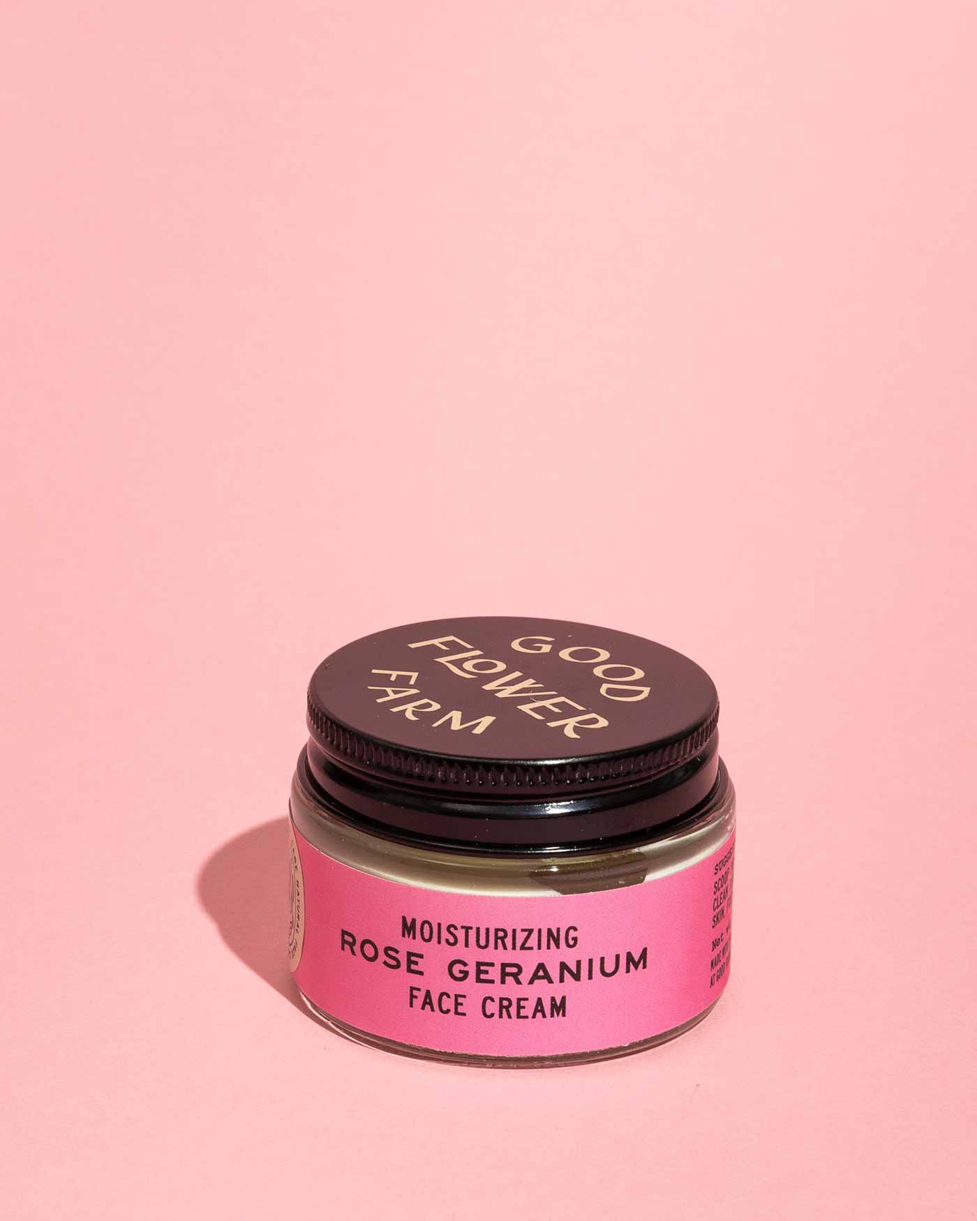 A small glass jar of Good Flower Farm Rose Geranium Face Cream with a black lid and pink label, shown against a solid light pink background.