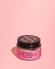 A small glass jar of Good Flower Farm Rose Geranium Face Cream with a black lid and pink label, shown against a solid light pink background.