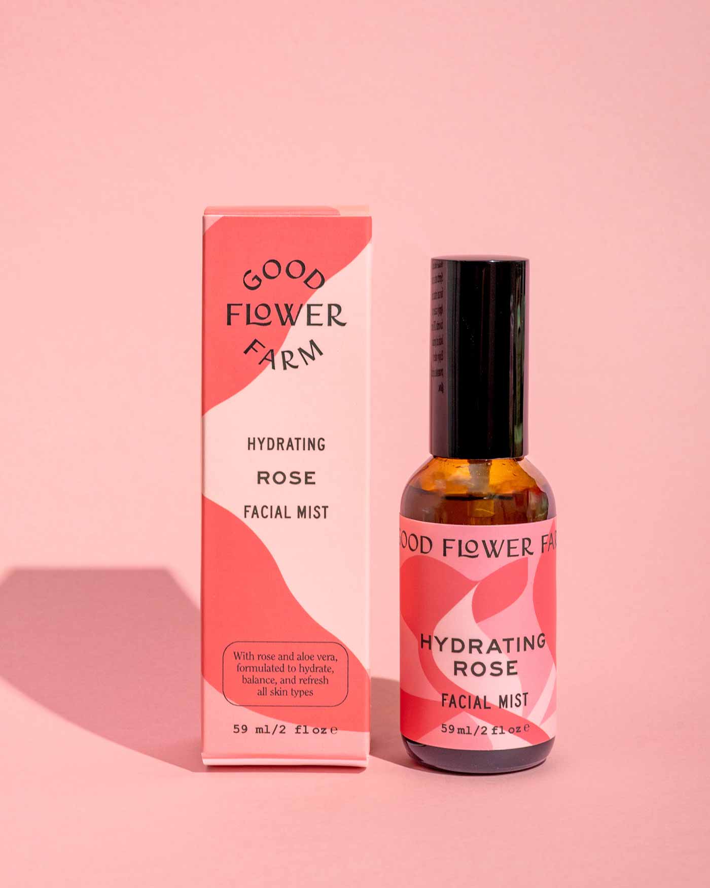 A bottle of Good Flower Farm Hydrating Rose Facial Mist, featuring bold modern patterns and black text on its pink and red box, is displayed on a pink background.