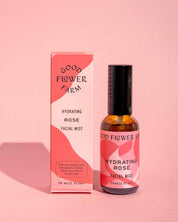 A bottle of Good Flower Farm Hydrating Rose Facial Mist, featuring bold modern patterns and black text on its pink and red box, is displayed on a pink background.