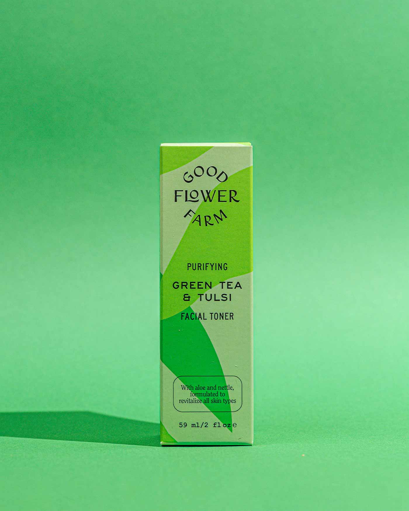 A box of Good Flower Farm Green Tea & Tulsi Facial Toner stands upright on a green surface with a matching background, showcasing its green tea, nettle ingredients, and revitalizing skin benefits.