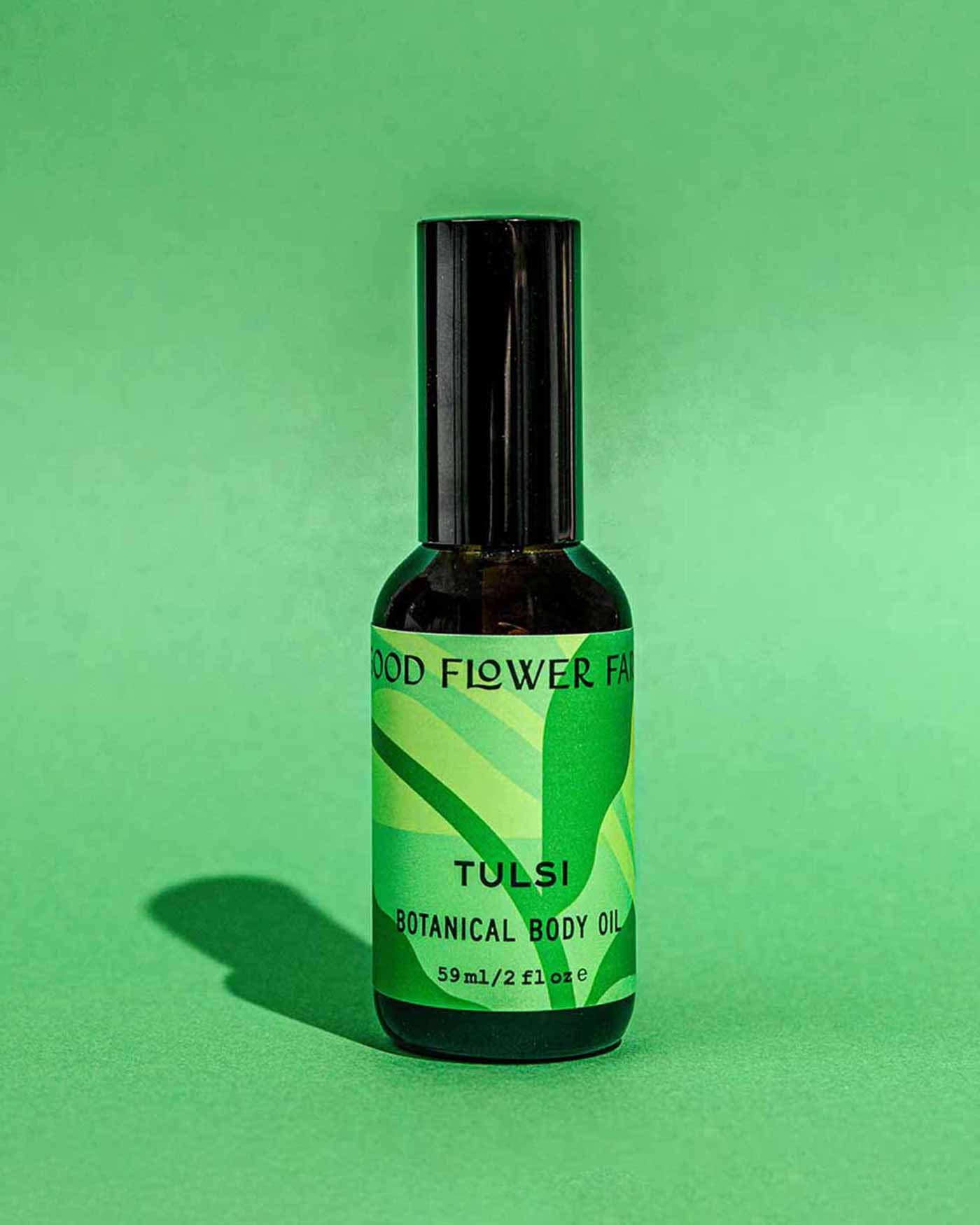 A dark bottle of Good Flower Farm’s TESTER — Tulsi Botanical Body Oil sits on a green background, with a bright green label featuring leaf graphics and text, its shadow reflecting the product’s Ayurvedic roots.