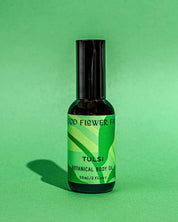 A dark bottle of Good Flower Farm’s TESTER — Tulsi Botanical Body Oil sits on a green background, with a bright green label featuring leaf graphics and text, its shadow reflecting the product’s Ayurvedic roots.