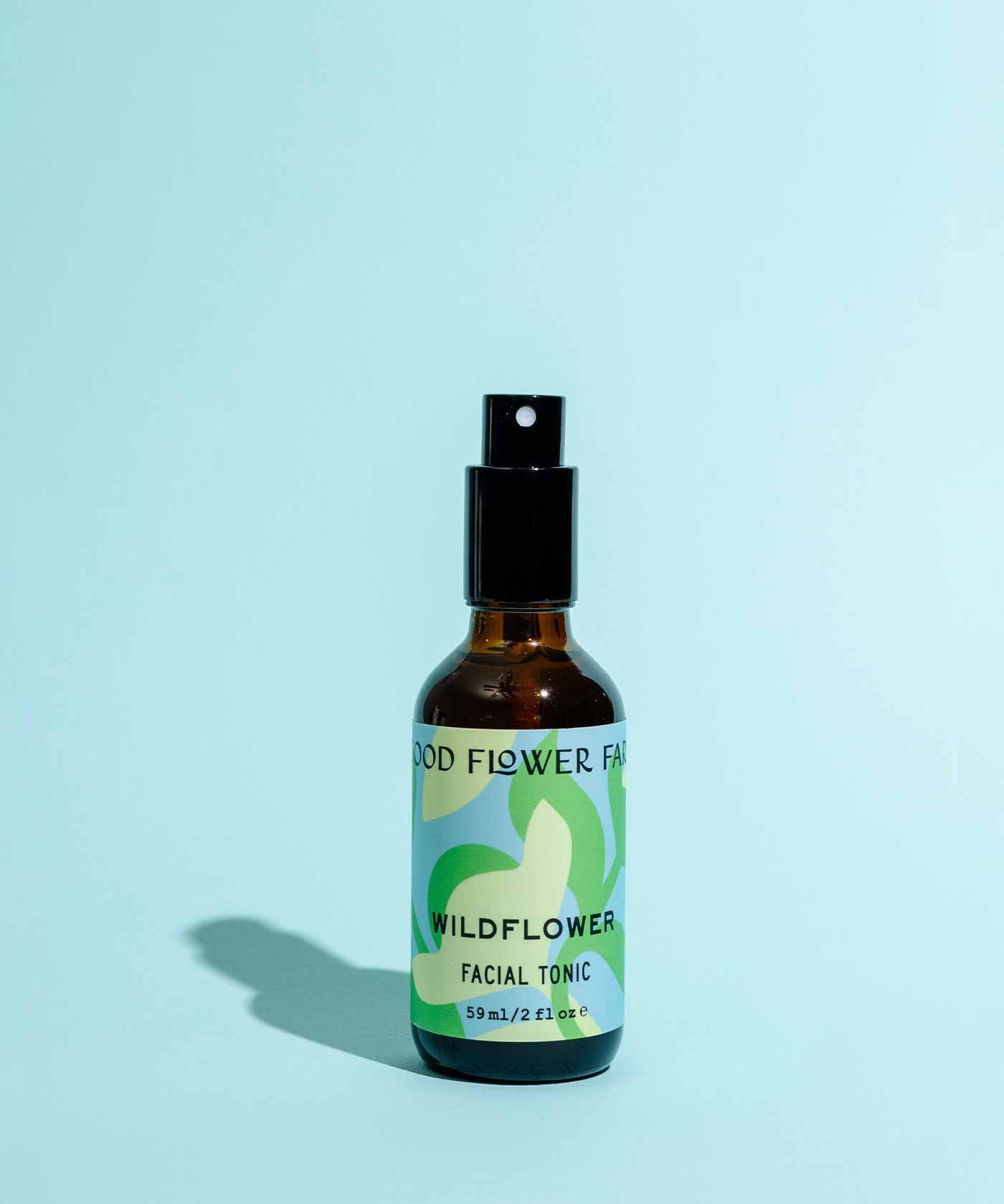 The Wildflower Facial Tonic by Good Flower Farm comes in a brown glass spray bottle with a green and white label, shown upright on a light blue background—formulated with wild herbal flowers to nourish your skin.