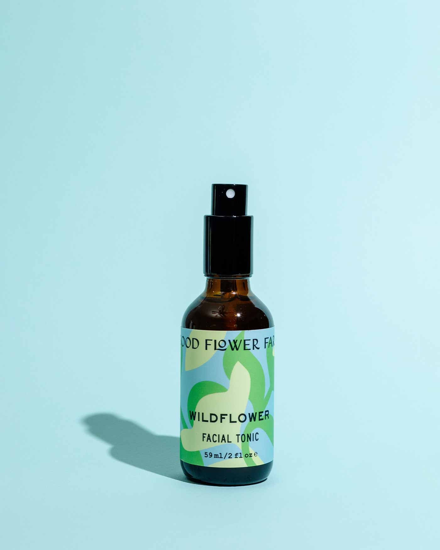 The Wildflower Facial Tonic by Good Flower Farm comes in a brown glass spray bottle with a green and white label, shown upright on a light blue background—formulated with wild herbal flowers to nourish your skin.