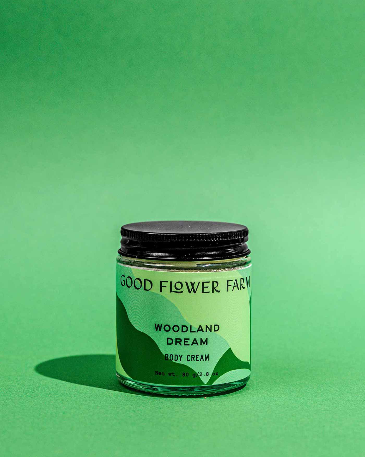 A jar of Good Flower Farm Woodland Dream Organic Body Cream rests on a green surface with a matching background. The light green label shows dark green accents and black text, and the jar is completed by a black lid.