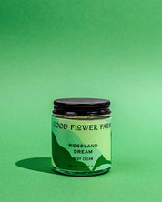 A jar of Good Flower Farm Woodland Dream Organic Body Cream rests on a green surface with a matching background. The light green label shows dark green accents and black text, and the jar is completed by a black lid.