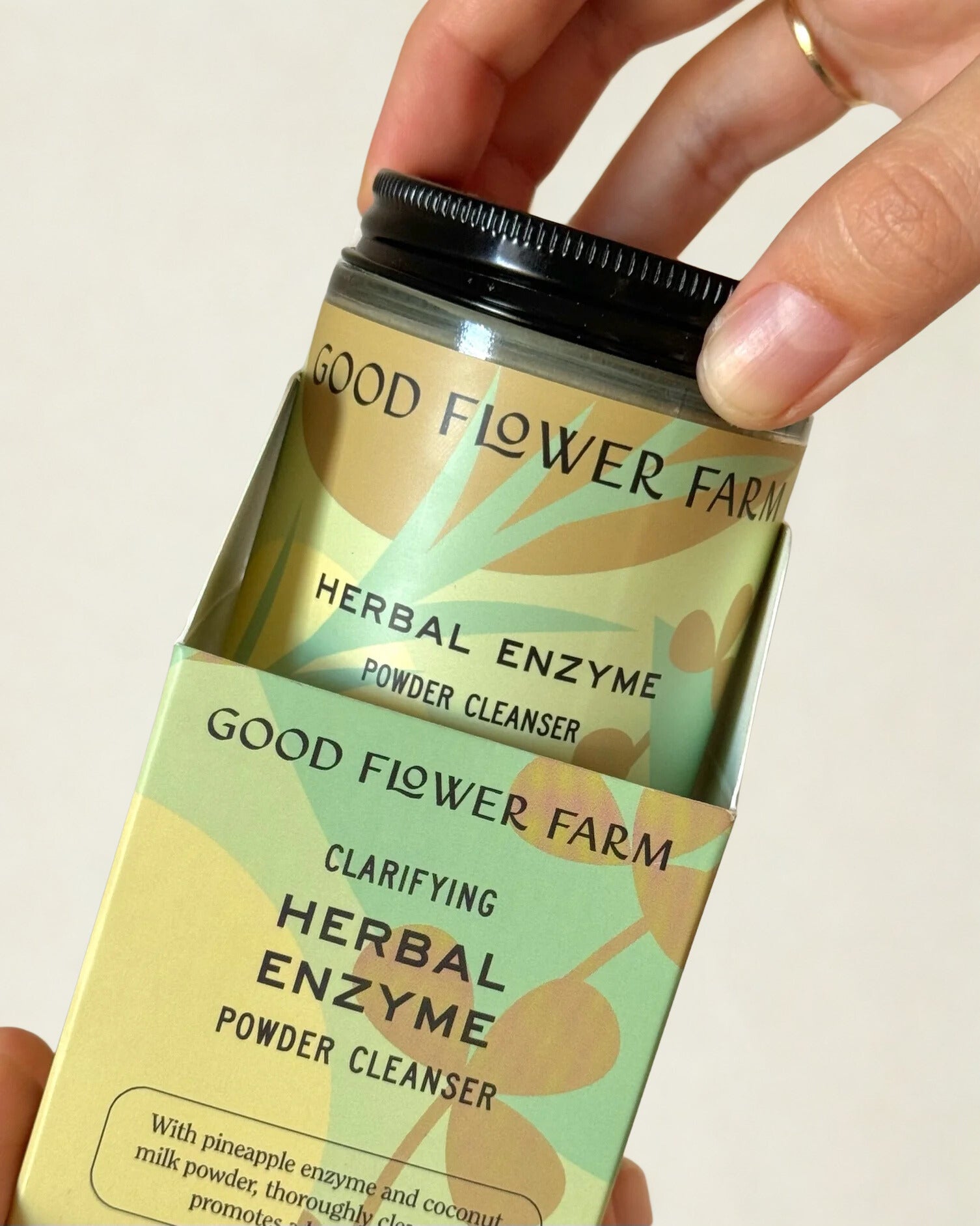 A hand takes a jar of Good Flower Farm Herbal Enzyme Powder Facial Cleanser from its green and yellow box, which showcases leafy designs and details about papaya enzymes on the vibrant packaging.