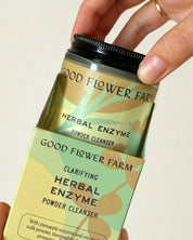 A hand takes a jar of Good Flower Farm Herbal Enzyme Powder Facial Cleanser from its green and yellow box, which showcases leafy designs and details about papaya enzymes on the vibrant packaging.